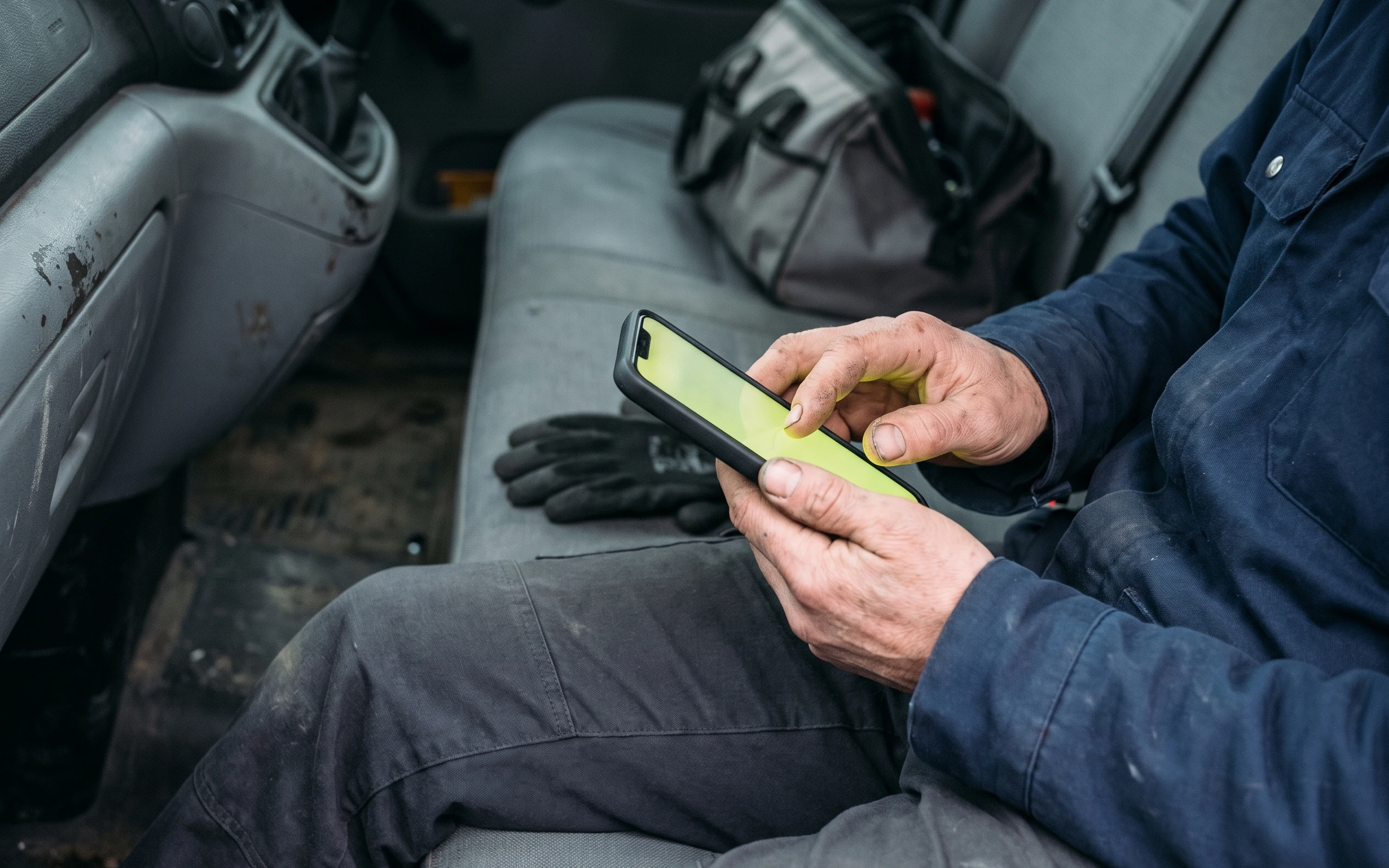 A contractor in work clothes answering the check on a phone from the seat of a van, the screen glowing chartreuse.